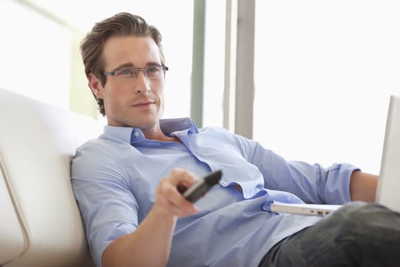 A man in glasses watches TV while browsing on his laptop.