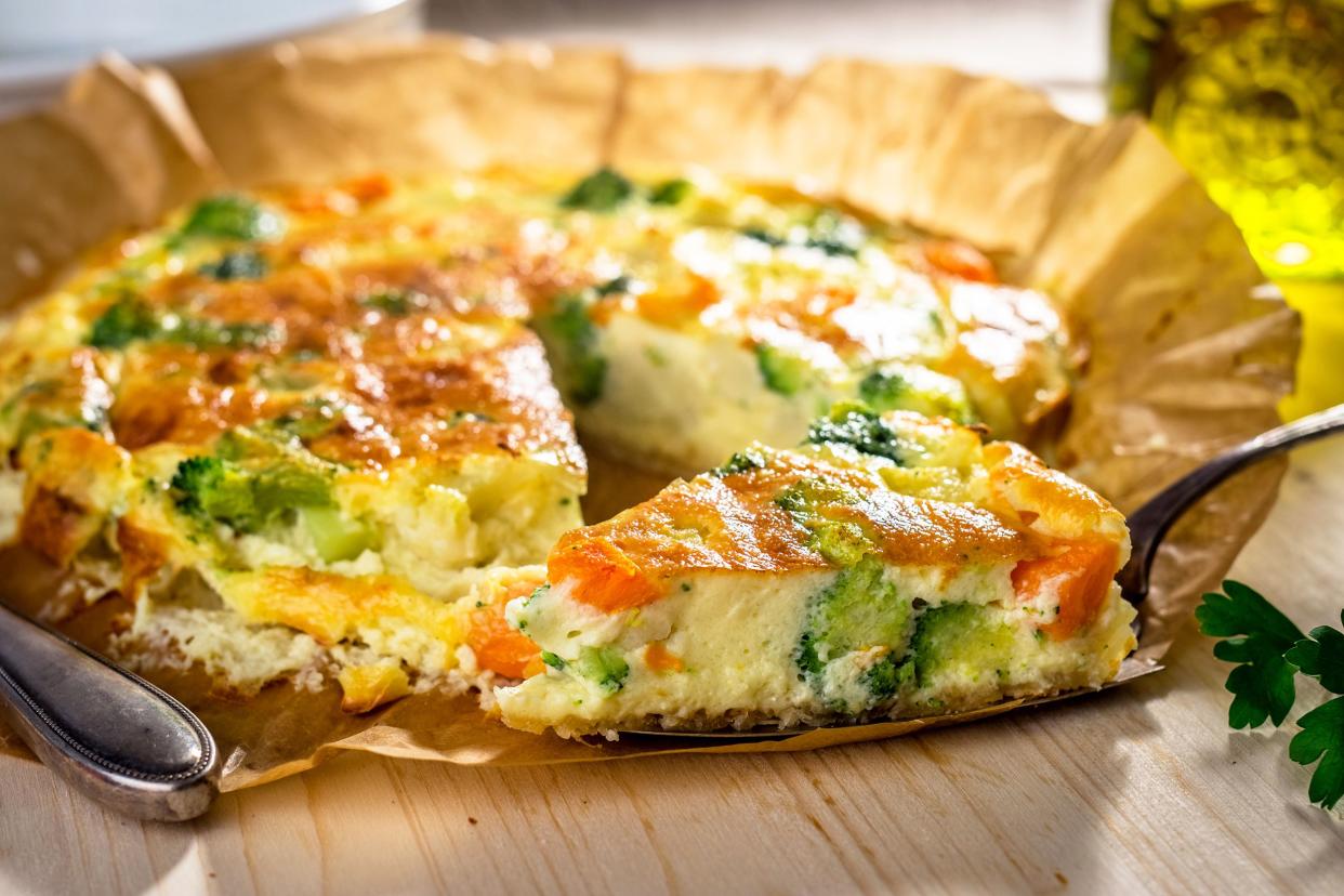 Slow-cooker crustless Mediterranean quiche on a piece of round brown parchment paper, one slice on a pie server, selective focus, sunlight coming from top right, on a wooden table, blurred