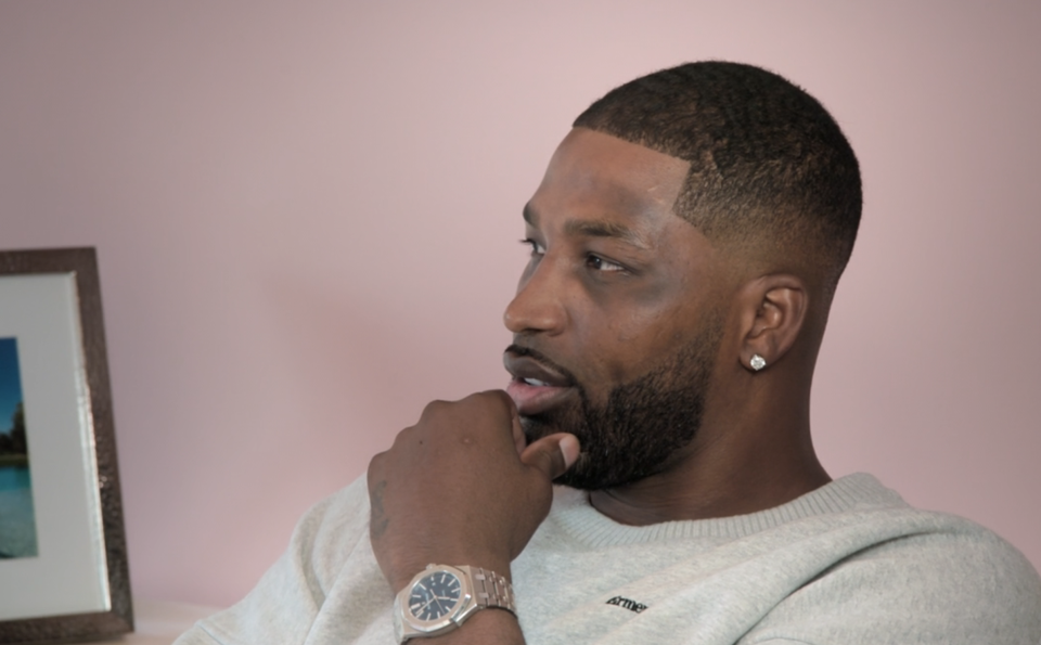 Tristan Thompson is sitting indoors, wearing a light sweater and a watch, with a thoughtful expression. A framed photograph and a smaller inset image are visible