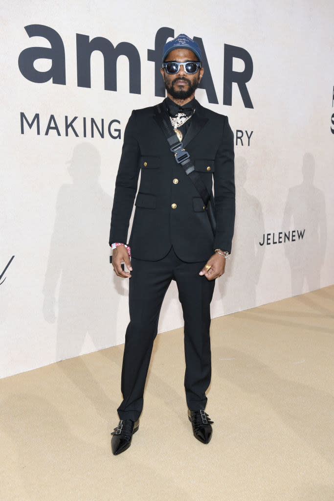 LaKeith in a tight suit with shoulder bag, hat, and sunglasses