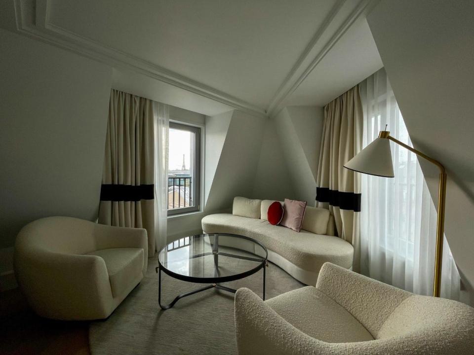 A view of the Eiffel Tower through a window in a room with a modern sofa, two chairs, and a coffee table.