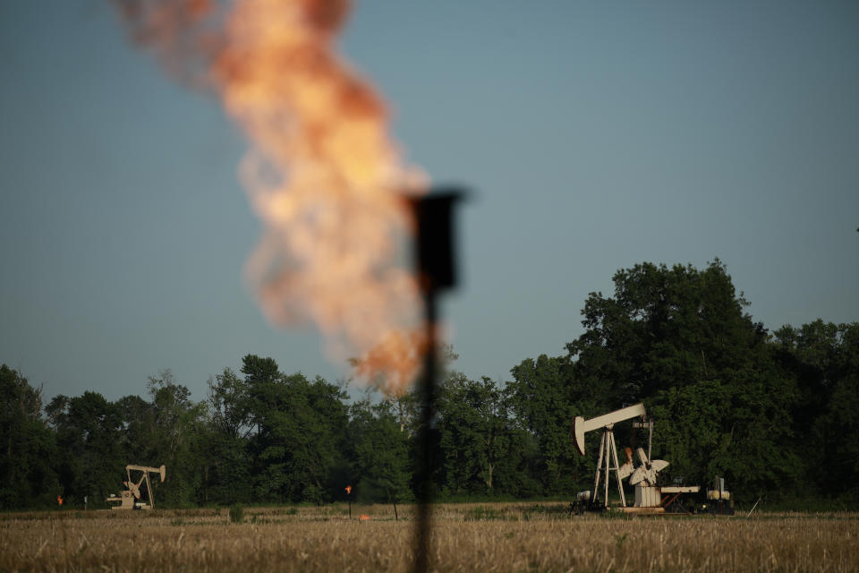 A natural gas flare burns near an oil pump jack in a field, with trees in the distance.