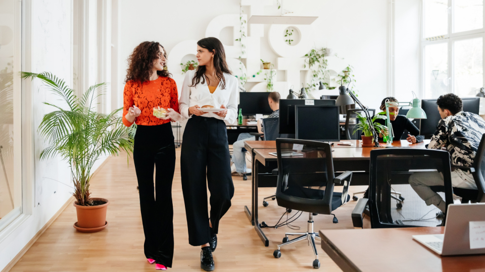 Two women walk through open plan office, talking. 