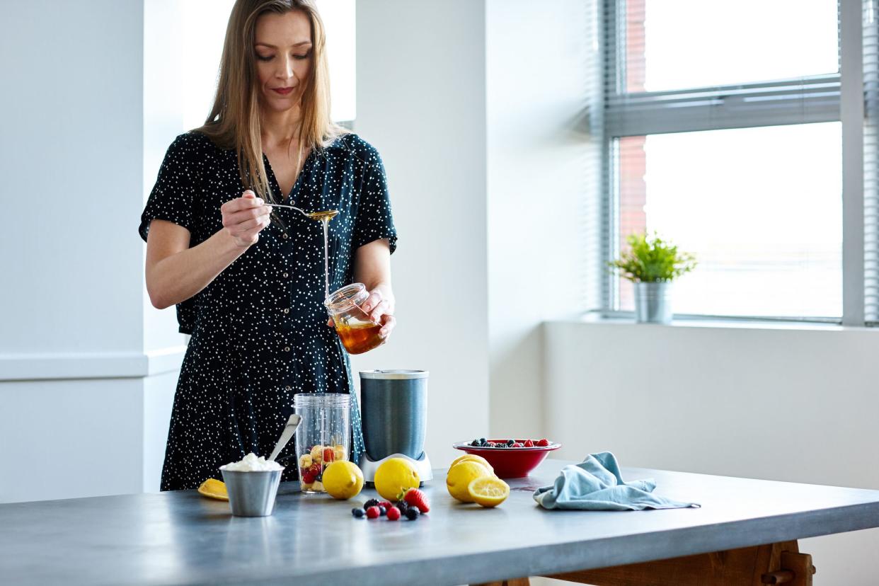 Beautiful mature woman adding honey in mixer jar with fruits. Woman preparing tropical beverage at home.