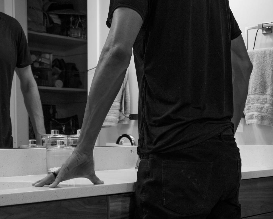 A black and white image of a man leaning on a bathroom counter and looking into a mirror, his head is not shown.