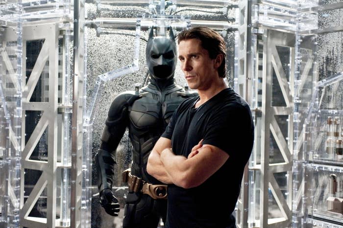 Christian Bale stands with arms crossed in front of a Batman suit display in a high-tech room in a scene from Batman Begins