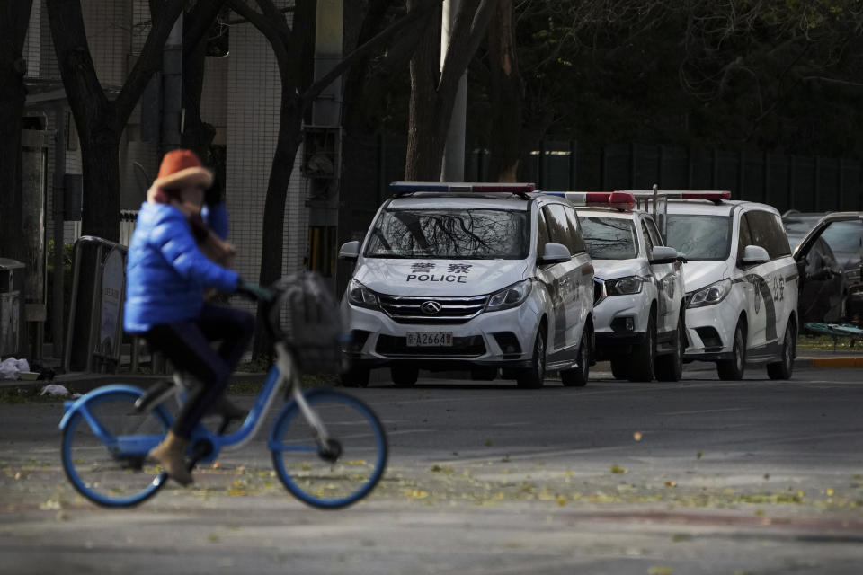 A woman rides past police vehicles parked along a road monitoring near the site of last weekend protest in Beijing, Tuesday, Nov. 29, 2022. With police out in force, there was no word of protests Tuesday in Beijing, Shanghai or other major cities. (AP Photo/Andy Wong)