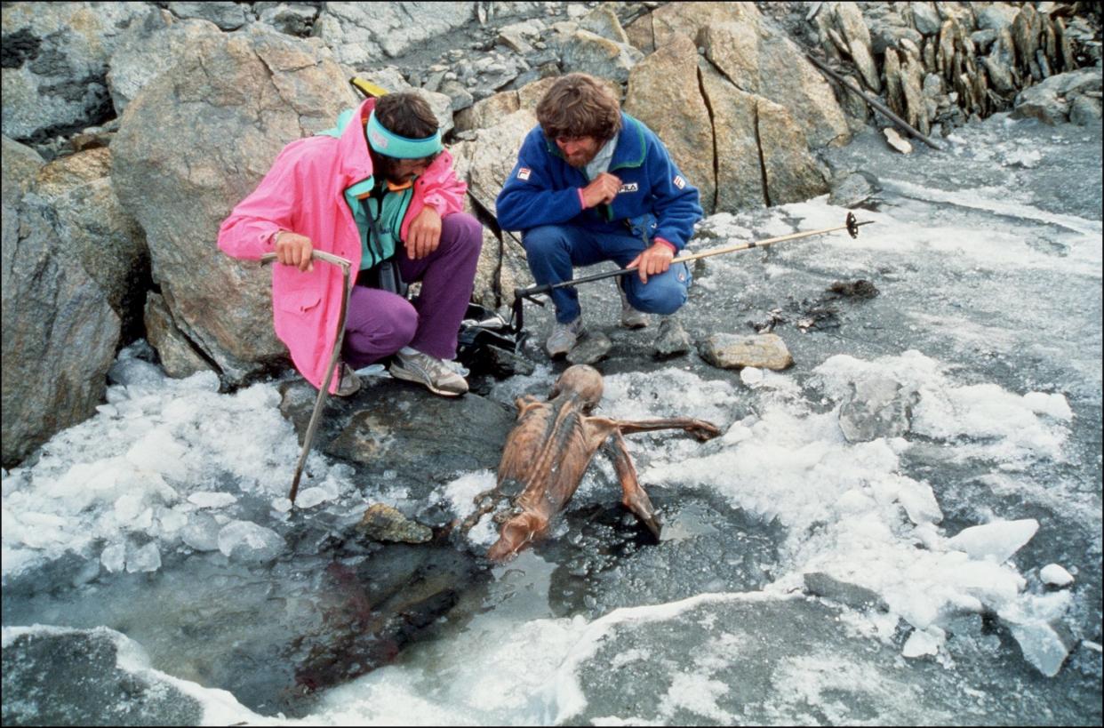 Two men with shaggy hair in '90s hiking clothes crouch on the ice beside a facedown mummy positioned as if it's crawling out of a puddle.