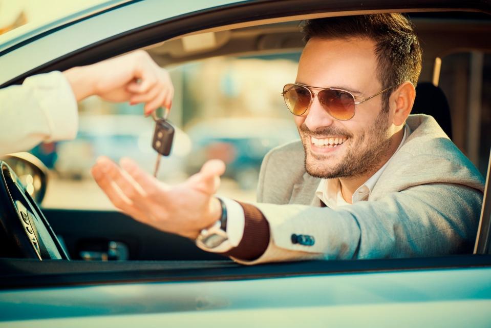 A smiling person sitting in his car and holding his hand out the window to take someone else's keys.