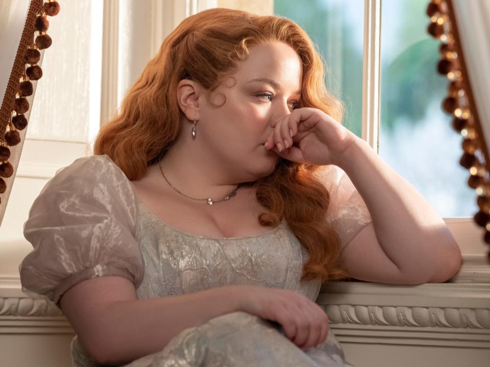 nicola coughlan as penelope featherington in season three of the show, wearing a blue and white shimmering dress, her hair styled in elegant, loose curls, looking forlornly out of a sunny window while seated on a chaise