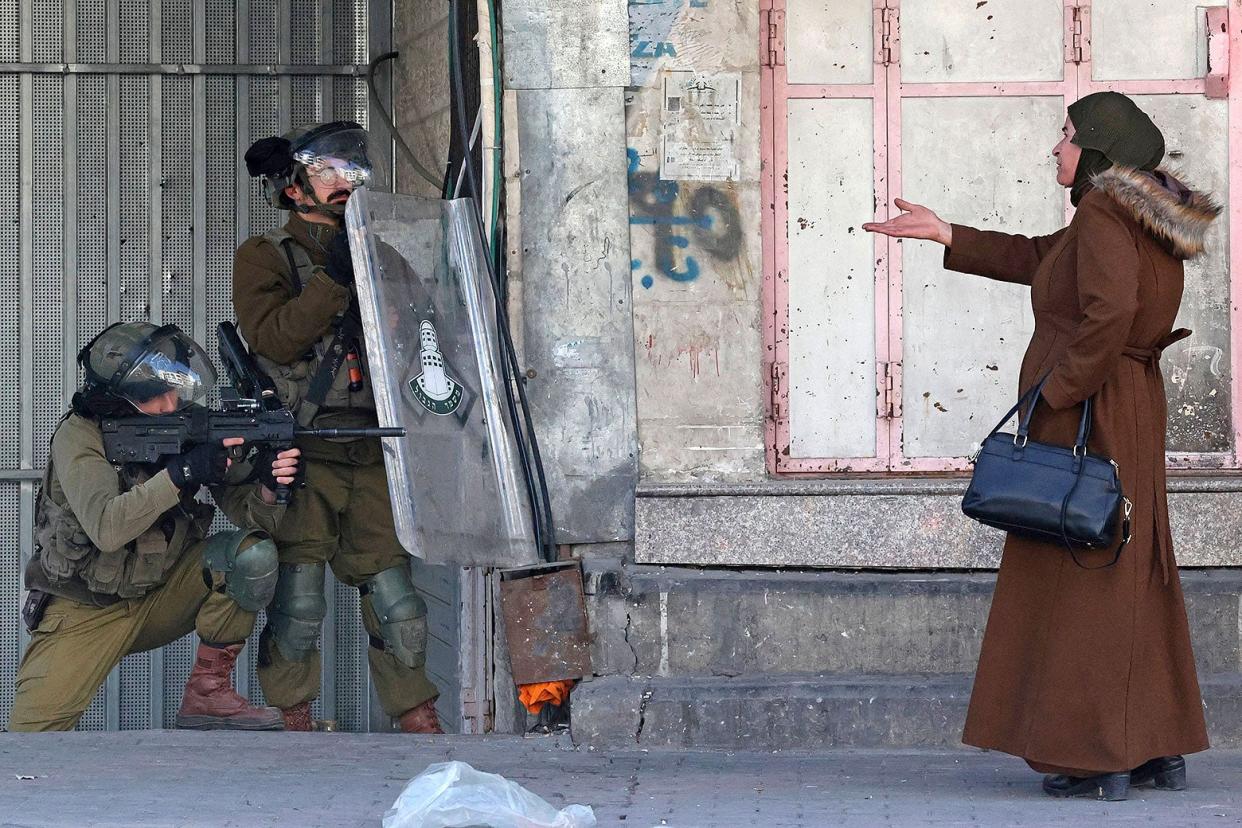 Two Israeli soldiers stand outside a shabby-looking building. A woman wearing a brown fur-lined coat holds a purse and gestures toward the soldiers with her right hand.