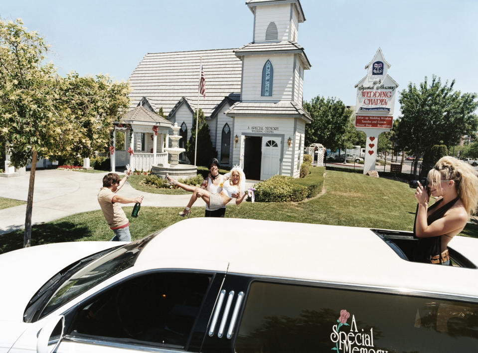 A bride playfully kicks her groom outside a wedding chapel as a photographer captures the moment; a limo is in the foreground