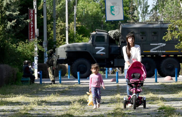 Esta foto tomada el 20 de mayo de 2022 muestra a una mujer y un niño caminando en un parque mientras los militares rusos patrullan la calle en Skadovsk, de Kherson