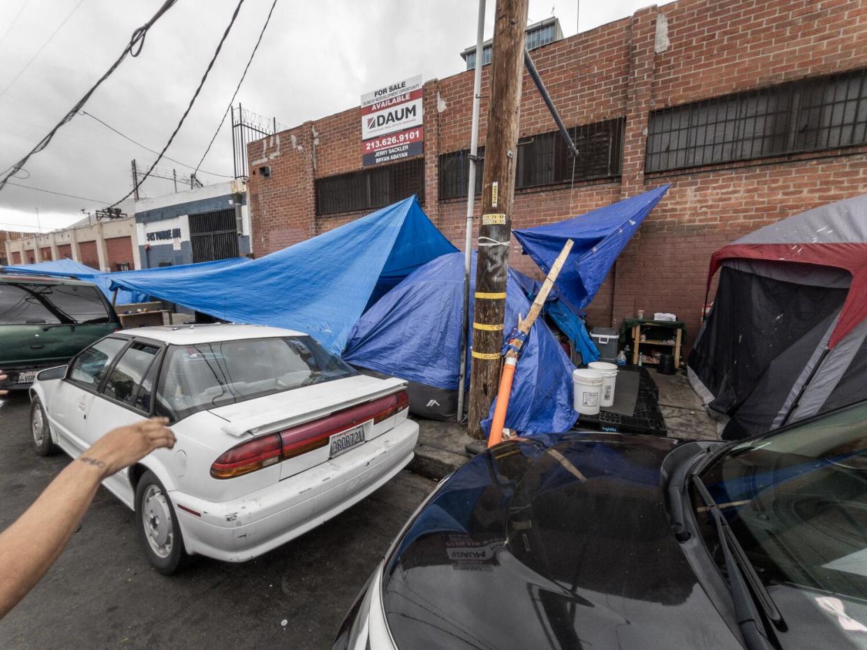 Tents and hanging tarps on a sidewalk next to parked cars.