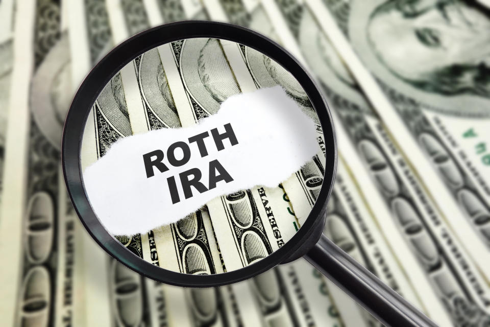 A magnifying glass over the words Roth IRA printed on a scrap of paper, on a bed of hundred-dollar bills.