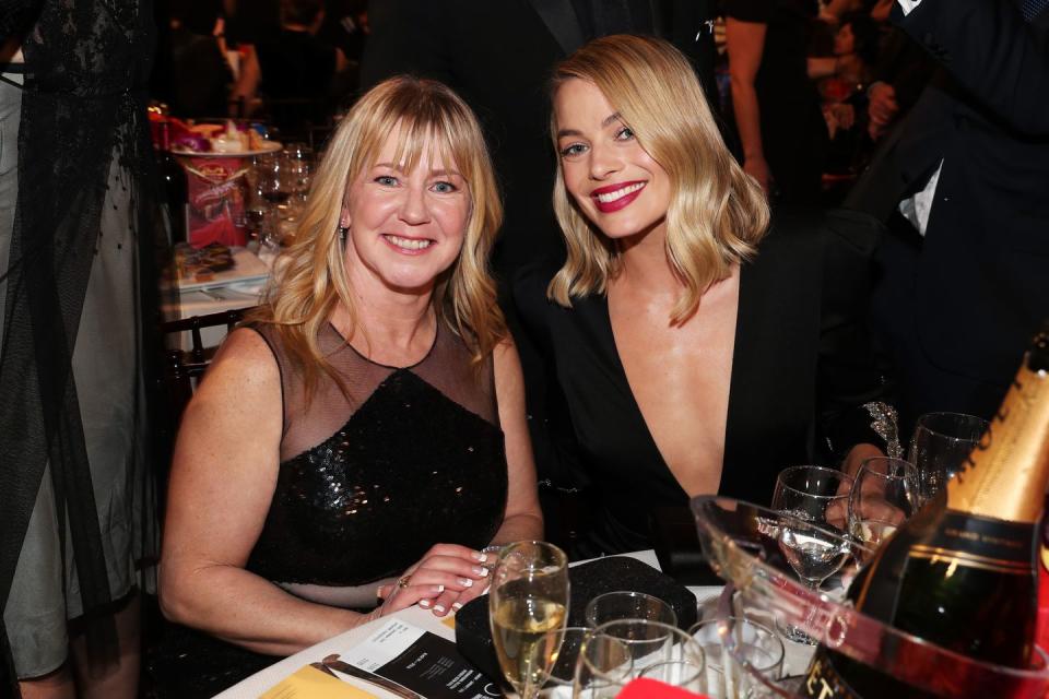 tonya harding and margot robbie smile at the camera while sitting at a table filled with glasses and other items, both women wear black dresses and have shoulder length blonde hair