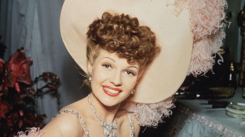 rita hayworth smiles at the camera, she wears a feathery top with bejeweled straps and a large matching hat as well as a necklace and earrings
