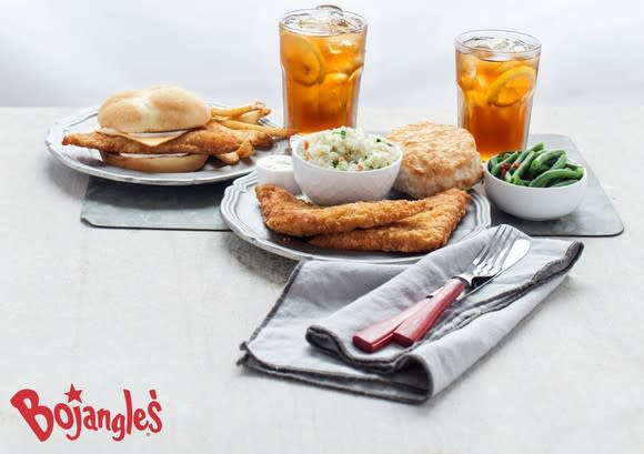 A BojAngler sandwich platter served on porcelain plates with two glasses of iced tea and two sides of veggies.