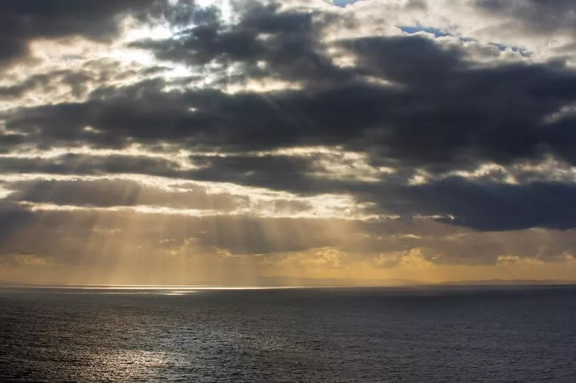 Sunrays bursting through dense sheet of rain clouds over sea water at sunset.