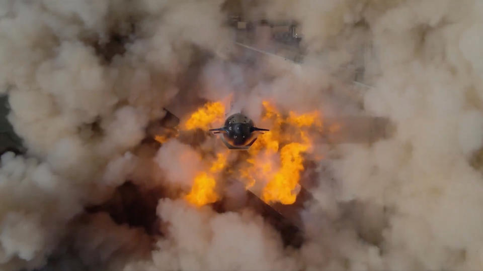 aerial view looking down on a rocket with flames erupting from its base and a dust cloud surrounding it.