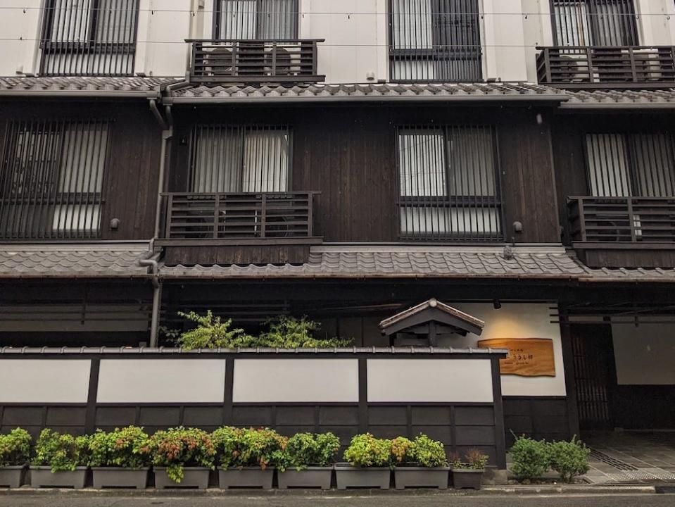 Image from the street of an off-white and brown building. There are rows of windows with small balconies, a tall fence on the ground level with an opening by the entrance, and planters in front of the fence with green bushes in them. Behind the fence is a wall with a wooden sign that has the hotel name written on it.