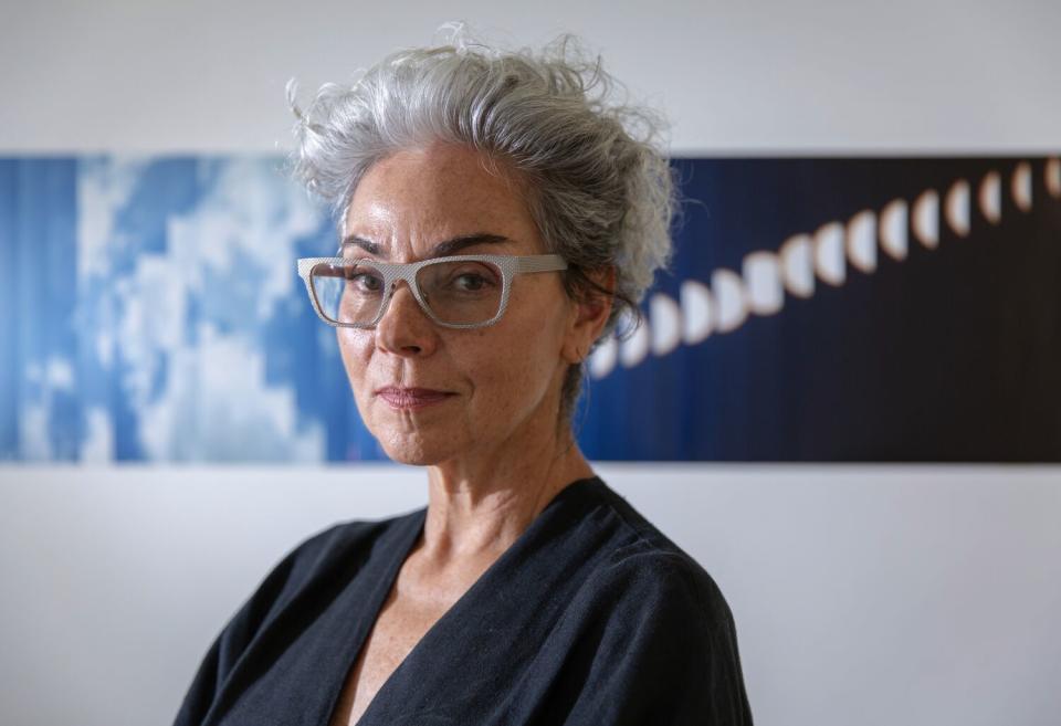 A woman in glasses and black shirt stands before a wall in her studio, which bears artwork depicting the sky.