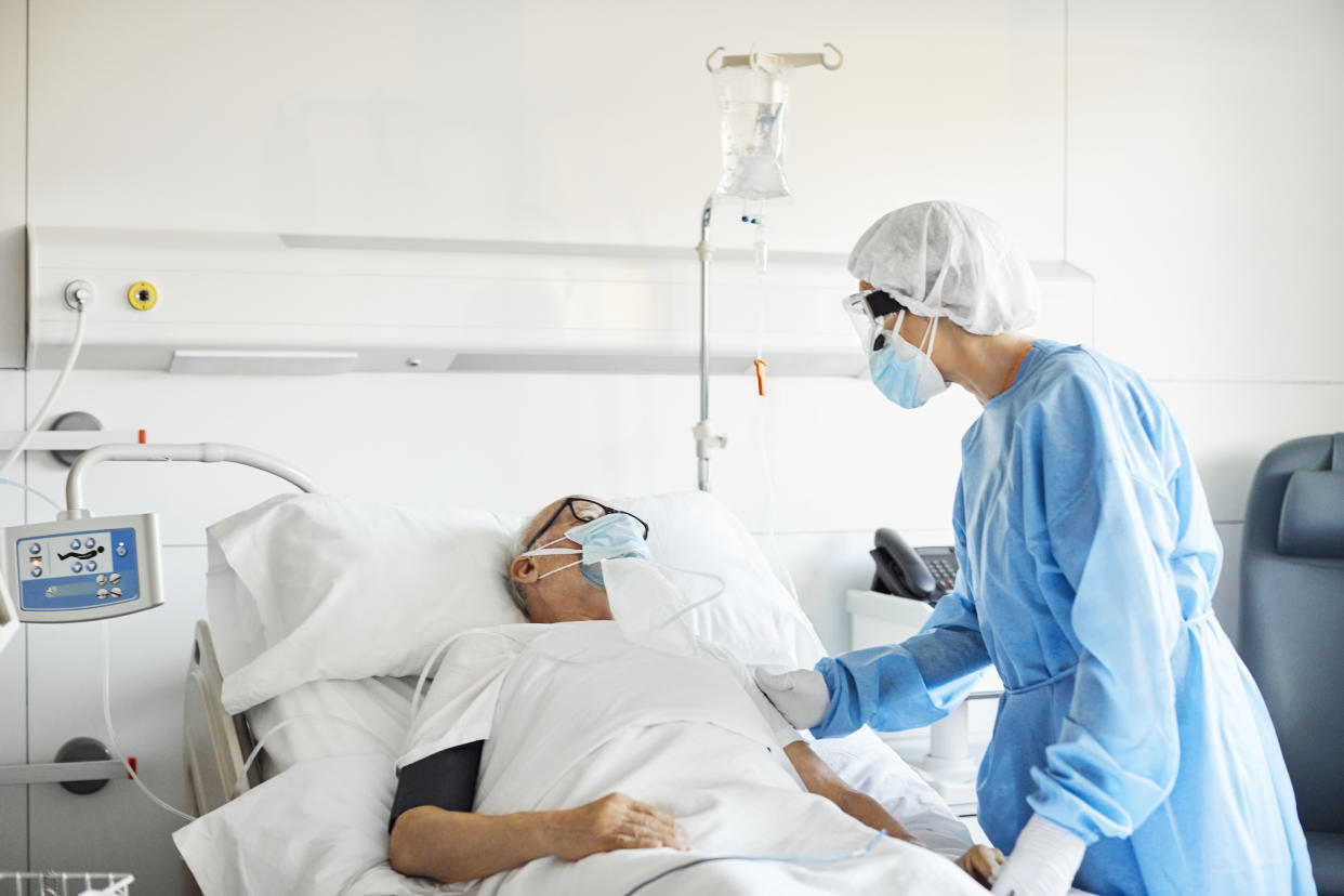 Female doctor in protective suit consoling senior patient. Elderly man with oxygen mask is lying on bed in intensive care unit during COVID-19 crisis. They are in hospital ward.