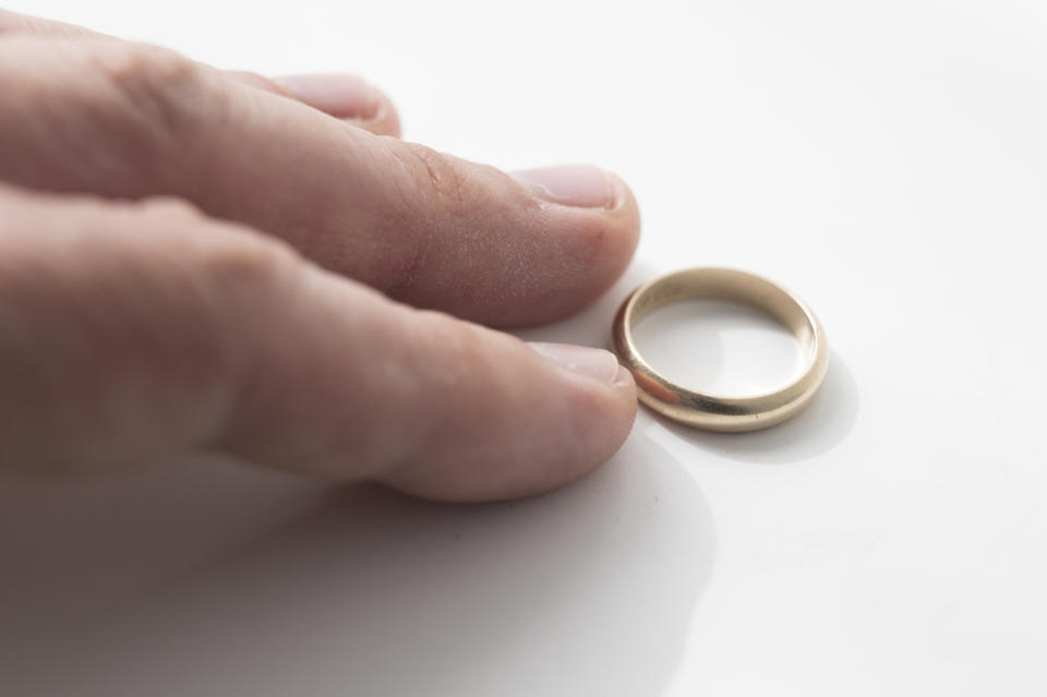 A hand gently touches a gold wedding ring lying on a white surface