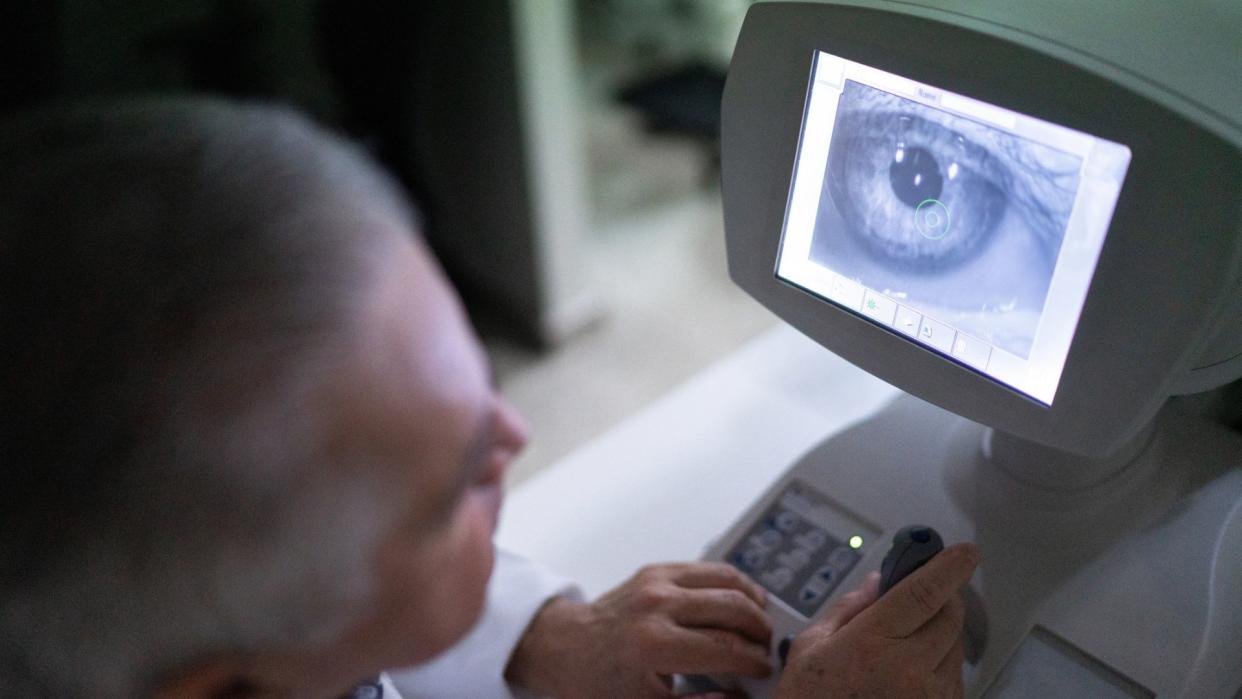  photo shows an eye doctor in the foreground, looking towards a computer monitor in the foreground that's showing a close up image of a patient's eyeball 