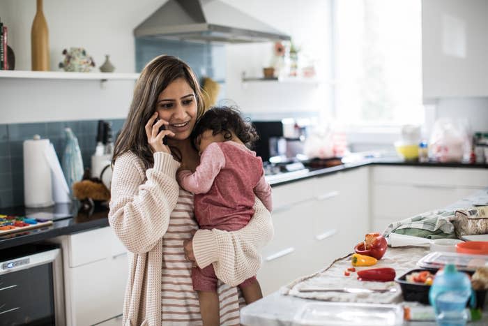 A woman stands in a kitchen holding a young child while talking on the phone