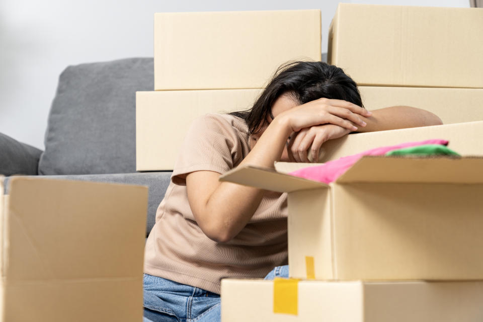 A person is sitting on the floor, leaning on cardboard boxes with their face buried in their arms, appearing stressed or tired