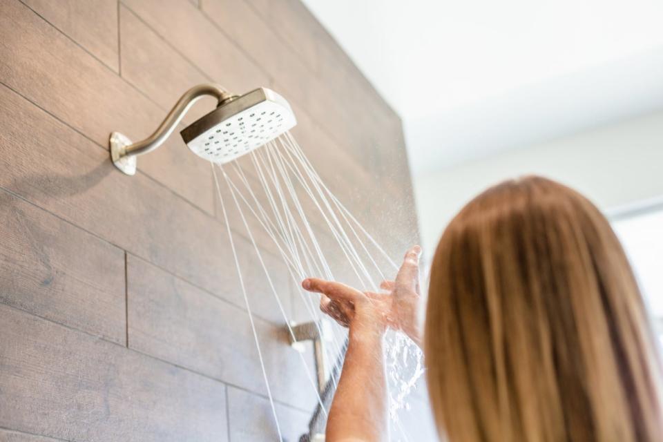 A woman runs her hands under the water streaming from the showerhead in the bathroom. 