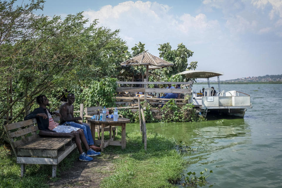 People rest on the mainland next to a floating restaurant and bar, on Lake Victoria near the Luzira area of Kampala, Uganda Sunday, Feb. 19, 2023. Flowering plants rise from the water into the wooden hull of James Kateeba's boat, used as a floating restaurant and bar that can be unmoored to drift for pleasure, but the greenery emerges from an innovative recycling project which uses thousands of dirt-encrusted plastic bottles to anchor the boat. (AP Photo/Hajarah Nalwadda)