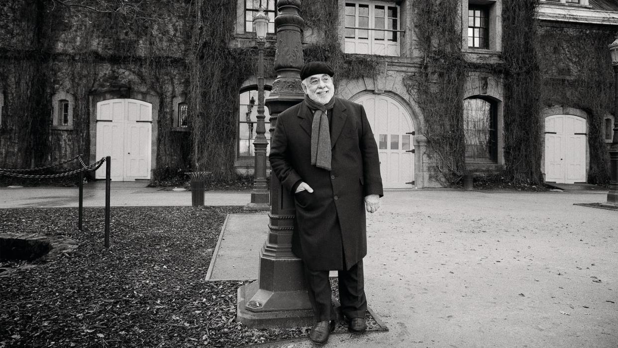 a bearded man in a suit with a beret and a scarf standing outside in fron t of a light pole