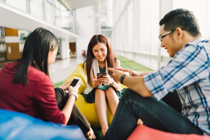 A group of smiling Asian youngsters sitting on bean bags while on their smartphones.