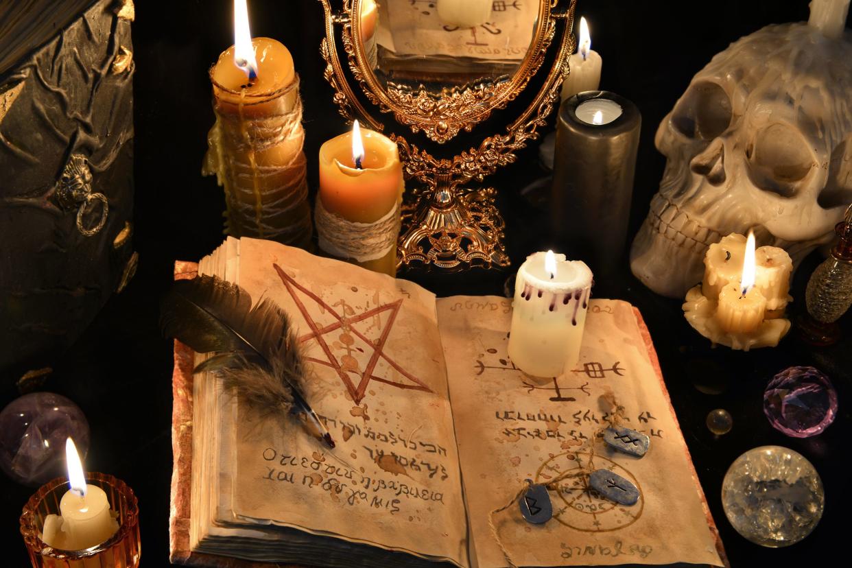 Wiccan altar with opened spell book surrounded by lit candles with a feather pen, a replica human skull, mirror, and crystal balls, in a dark room