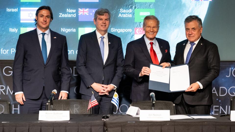 Four men in suits smile and hold up a signed document for the cameras to see