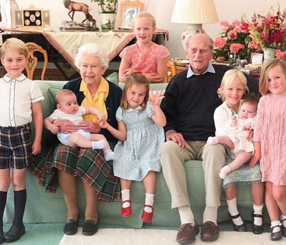 The Queen holding one of her great-grandchildren, sitting next to her late husband on a couch, surrounded by other great-grandchildren. 