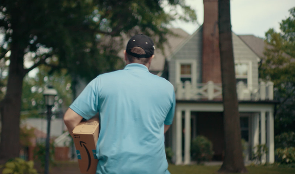 Man in light blue shirt carrying an Amazon package and walking toward a gray house.