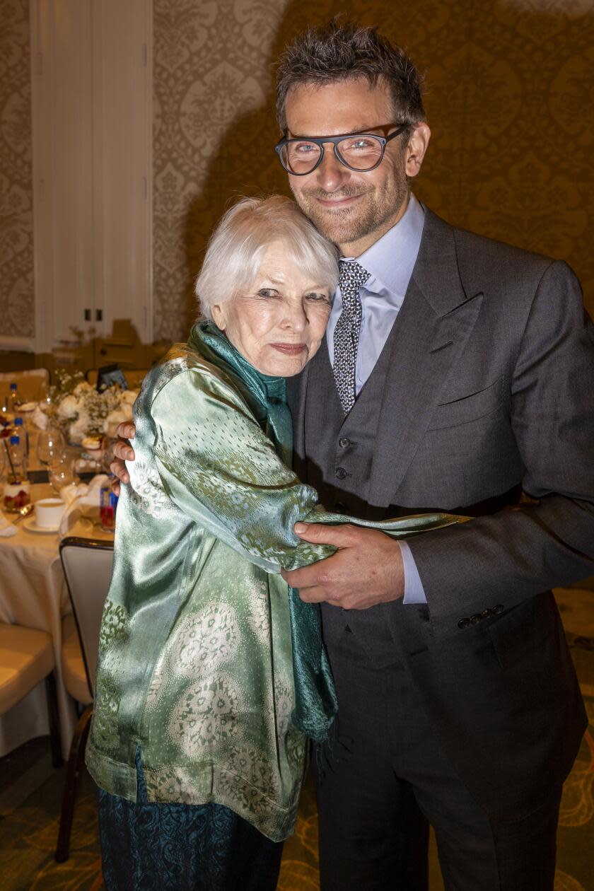 A gray-haired woman hugs a man in a suit around his waist