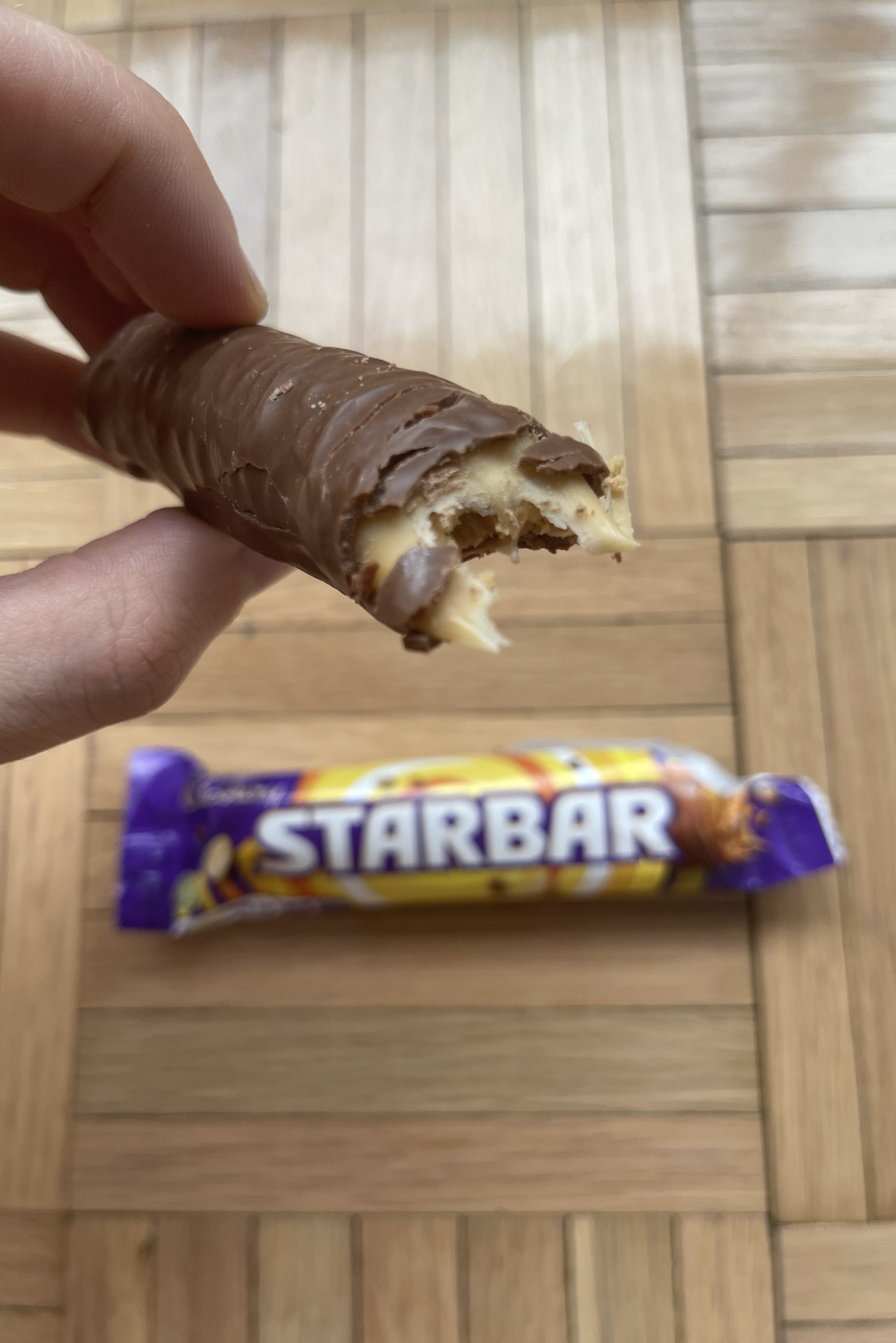 Hand holding a half-eaten Starbar chocolate with its wrapper on a wooden floor