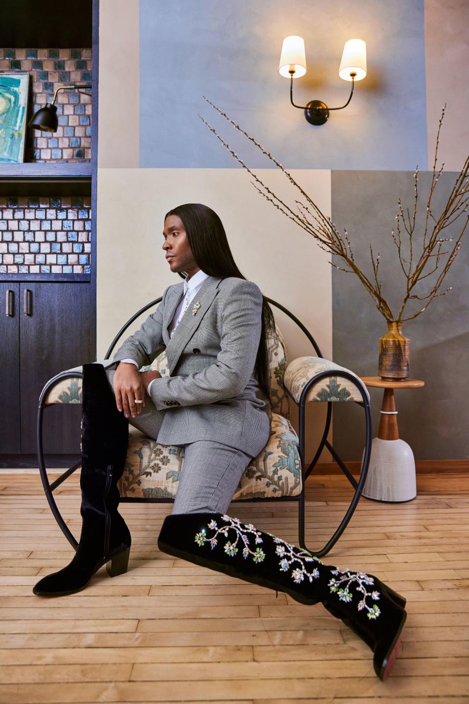 Law Roach sitting profile in a chair, long straight hair, gray suit, knee-high black embroidered boots