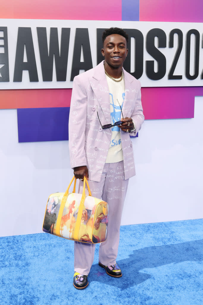 Olly Sholotan in a patterned suit and printed T-shirt holds colorful duffle bag and sunglasses at an awards show
