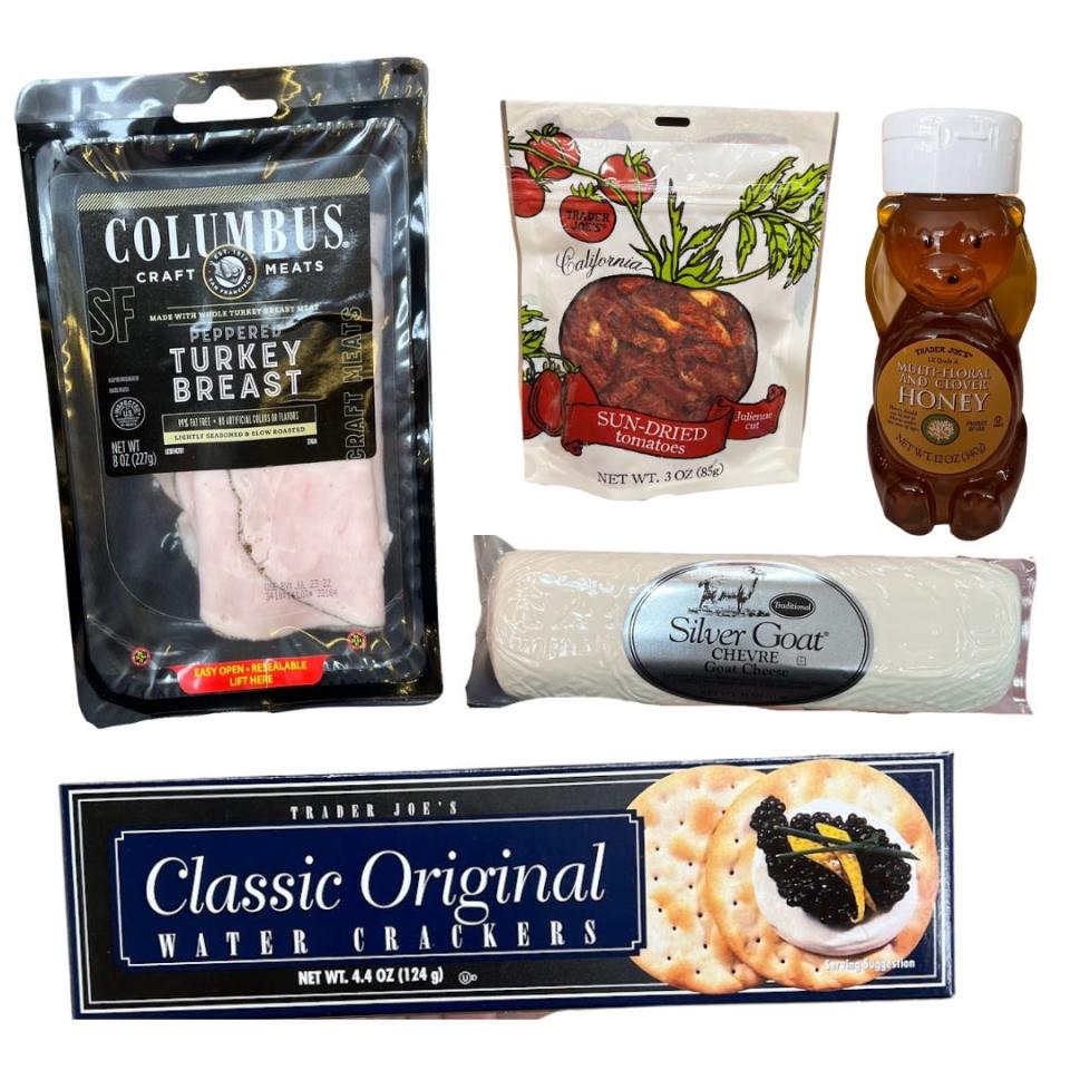 A black package of sliced turkey meat, a package of sun-dried tomatoes, a honey bear, a package of goat cheese, and a black box of crackers