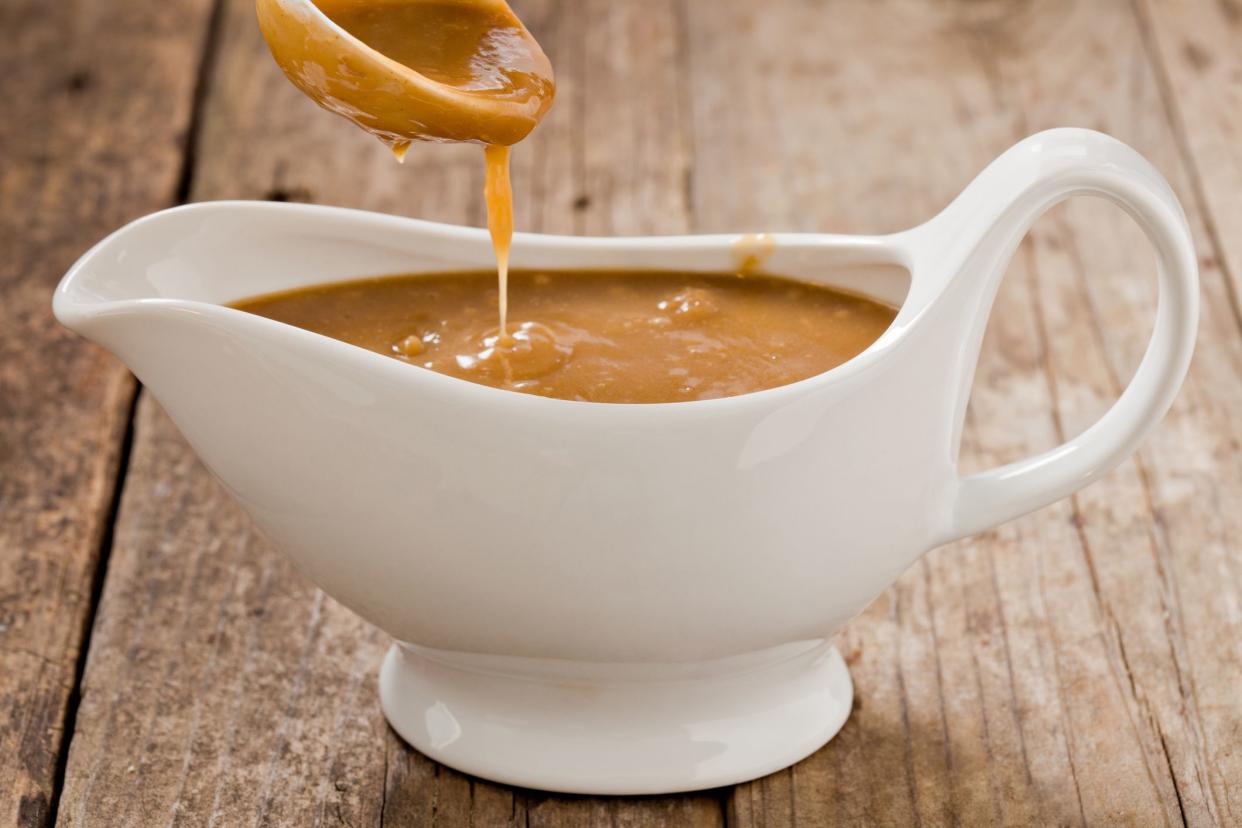 A close up shot of a small white ladle pouring brown gravy into a white ceramic gravy boat. Shot on a grungy old wooden table.