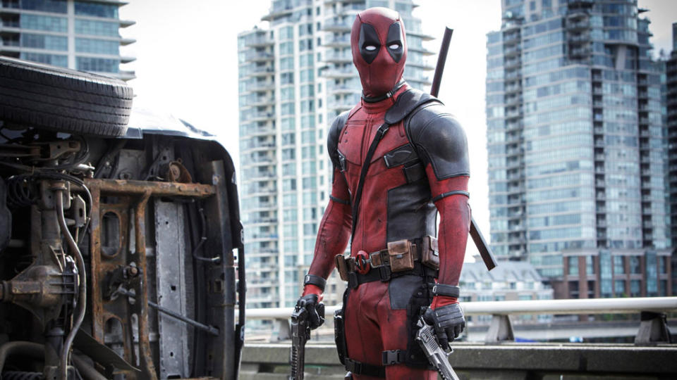 Deadpool holding a pair of guns as he stands next to an overturned vehicle.