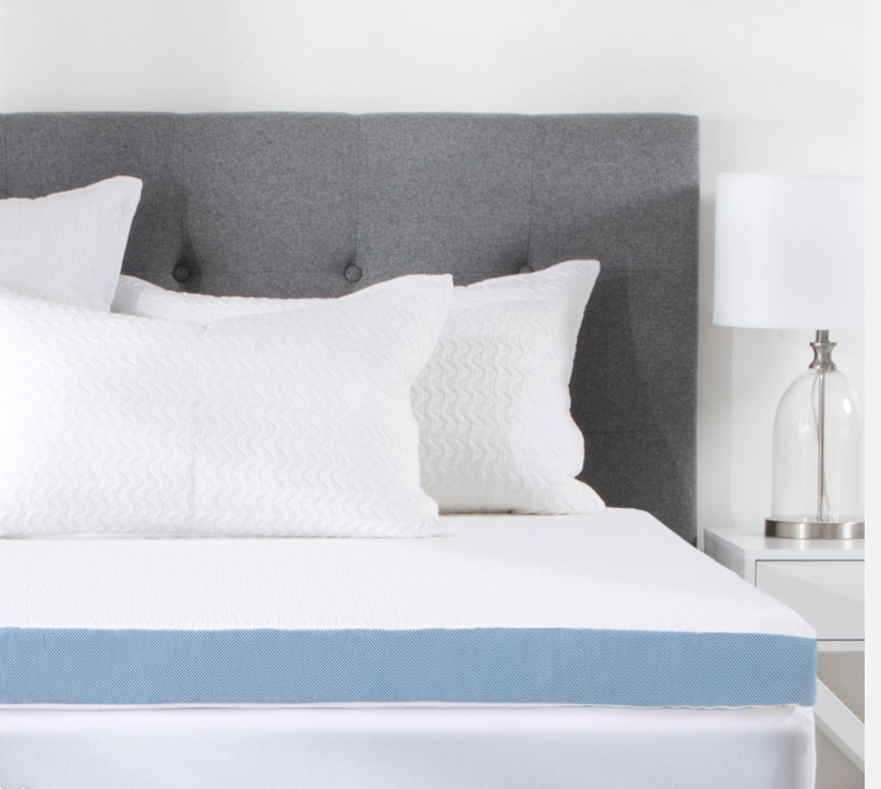 A white Hilton Relax Therapy 10cm Memory Foam Mattress Topper sits on a white mattress with white pillows and a grey fabric bed board, with lamp on a bedside table.