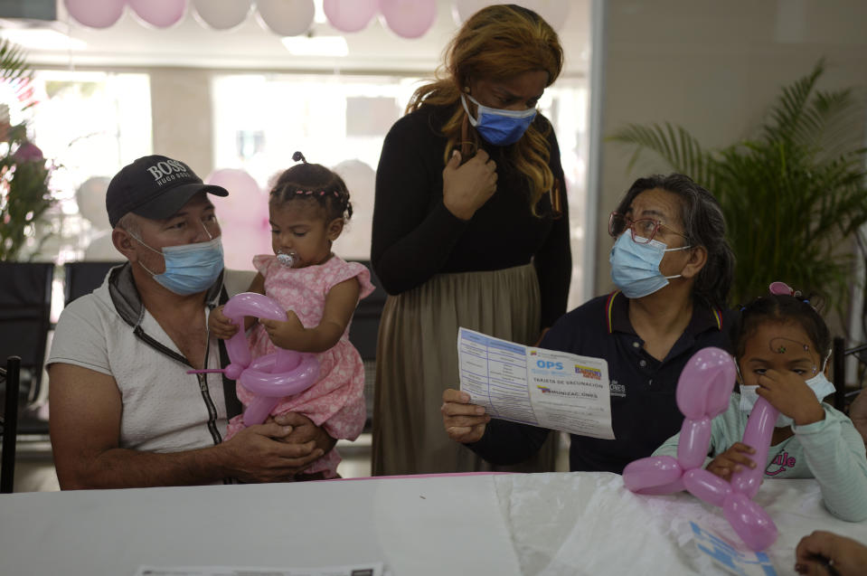 Dimas Campos sostiene a su hija Anastasia, de 19 meses, en sus brazos mientras su esposa (de pie) habla con una enfermera, que sostiene otra niña, sobre el historial de vacunaciones de su hija durante una campaña de vacunaciones gratis contra el polio, la rubeola y la influenza organizada por el Ministerio de Salud en Caracas el 18 de junio del 2022. (AP Photo/Ariana Cubillos)