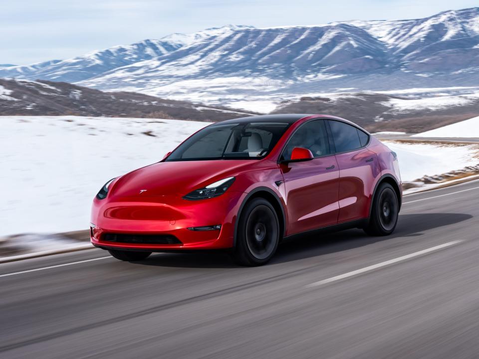 A red Tesla Model Y SUV drives down a road with snow and mountains in the background.