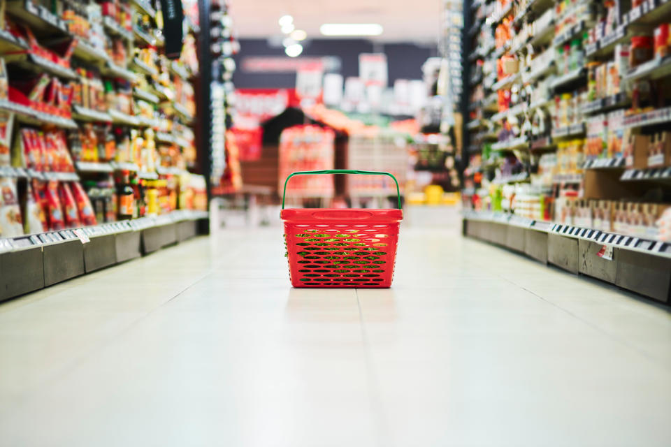 A red shopping basket sits in the middle of a grocery store aisle, surrounded by shelves stocked with various products